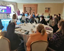 Room with round tables of people sitting looking at presentation on a screen.